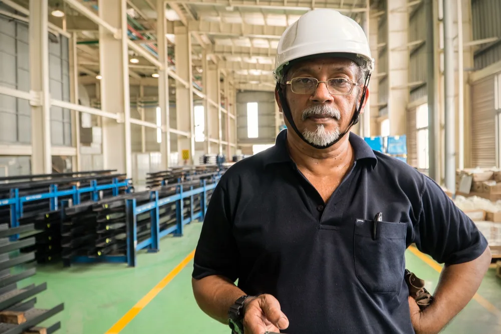 Portrait of a supervisor in a white hard hat on a factory floor, steel racking and the length of the shed receding behind him